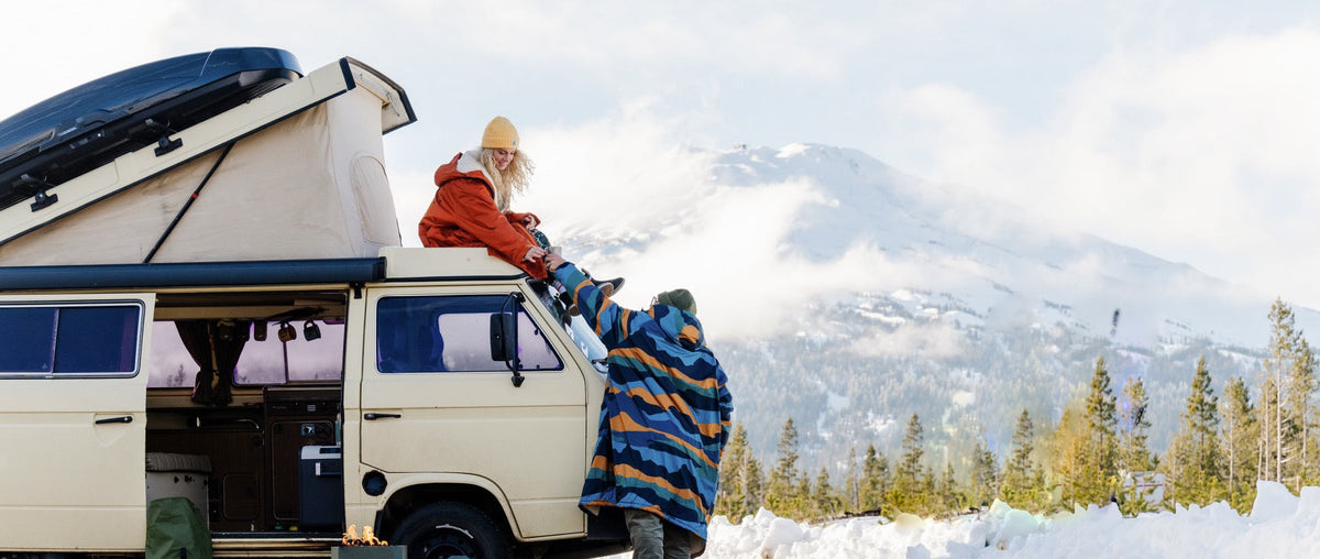 A woman sits on top of a retro camper and a man hands her a cup of coffee.