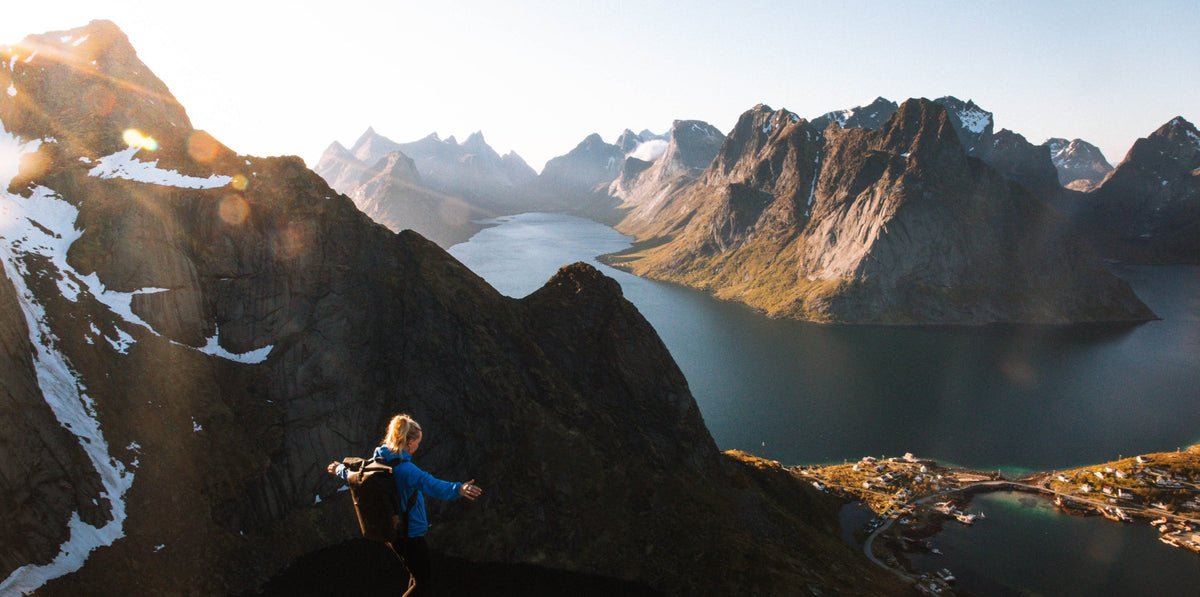A woman standing on a mountain with a fjord and snow capped peaks in the background